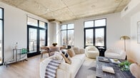 This thoughtfully designed one-bedroom, one-bathroom unit blends industrial and contemporary aesthetics into a cohesive, light-filled living space. The open-plan living and kitchen area is the architectural centerpiece, distinguished by a striking exposed concrete ceiling fitted with track lighting and an abundance of floor-to-ceiling black-framed windows and French doors that flood the interior with natural light and offer expansive outdoor views. Light hardwood flooring runs throughout, complementing the clean white walls and reinforcing the modern aesthetic.

The kitchen is outfitted with high-gloss white cabinetry, stainless steel appliances, and a generous island with bar seating, all illuminated by pendant lighting. A dedicated dining area sits adjacent, benefiting directly from the expansive window views. The living area offers generous proportions for comfortable furnishing arrangements.

The bedroom is a serene retreat with an exposed concrete ceiling detail, a large window, and ample built-in closet storage. A clean hallway with recessed lighting connects the private areas of the unit. The bathroom features large-format tile, a frameless glass-enclosed walk-in shower, and a warm wood-toned vanity, delivering a refined, spa-like finish throughout.