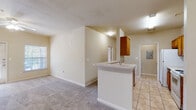 This single-story residence is thoughtfully laid out to maximize functionality and comfort throughout. The kitchen is designed in a classic galley style, featuring warm honey-toned wood cabinetry lining both walls, light countertops, and a full suite of white appliances including a coil-top range, built-in microwave, refrigerator, and dishwasher. Light tile flooring runs through the kitchen and extends toward a utility area, which houses an in-unit washer with overhead shelving for added storage convenience. A peninsula counter with a sink connects the kitchen to the open living area, creating a seamless flow between spaces. The living area is finished with neutral carpet and benefits from natural light filtering in through windows. A long hallway leads to the bedrooms, each featuring plush carpet, ceiling fans with light fixtures, and windows that invite natural light. The bathroom is well-appointed with warm wood vanity cabinetry, a wide countertop with dual sinks, a tiled tub-shower combination, and terracotta-toned tile flooring. Neutral wall tones throughout the residence create a clean, cohesive aesthetic that provides a versatile backdrop for a variety of interior styles.