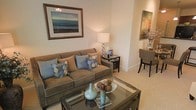 This single-floor space offers a thoughtfully designed interior that balances comfort and sophistication. The living area serves as the heart of the space, featuring a plush taupe sofa adorned with coordinated blue and patterned throw pillows that add a touch of personality to the neutral palette. A glass-topped coffee table with a metallic frame anchors the seating area, styled with decorative vases and natural accents that enhance the warm, inviting atmosphere. Soft, neutral wall tones and light carpeting throughout contribute to an overall sense of calm and cohesion. A framed landscape artwork above the sofa draws the eye upward, while a classic table lamp on a dark wood side table provides warm ambient lighting. The space transitions naturally into a dining area, visible in the background, where dark wood chairs and a compact table are accented with a floral arrangement and wall art, maintaining the refined aesthetic established in the living area. The overall design reflects a carefully curated, model-quality interior that emphasizes livability, coordinated furnishings, and a welcoming ambiance suited to everyday comfort and relaxed entertaining.