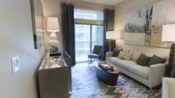 This single-floor living space showcases a thoughtfully designed interior that balances comfort and contemporary style. The main living area serves as the centerpiece of the space, featuring a light gray sofa adorned with patterned accent pillows, complemented by a sleek, rounded dark coffee table that anchors the seating arrangement. A boldly patterned area rug adds texture and visual interest to the floor beneath.

Natural light floods the room through a large sliding glass door dressed with floor-to-ceiling charcoal drape panels, which open directly onto a private balcony. The balcony, partially visible through the glass, offers an outdoor retreat complete with a tufted accent chair, ideal for relaxing while enjoying views of the surrounding urban landscape.

Interior lighting is carefully layered, with tall white ceramic table lamps providing warm ambient illumination alongside a ceiling fan overhead. A mirrored console table near the entrance adds depth and brightness to the space. Abstract wall art in cool tones contributes a refined artistic touch. The overall ambiance is polished and inviting, reflecting a modern urban aesthetic throughout.