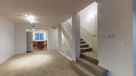 This two-story residence offers a well-organized and functional layout spread across two floors. The main level is anchored by a spacious carpeted living area that provides a warm and comfortable atmosphere, with neutral tones throughout that lend a clean, cohesive aesthetic. A centrally positioned staircase with classic white balusters and a sturdy handrail connects the two levels, serving as both a practical and visually defining architectural element.

Toward the rear of the main floor, the kitchen is thoughtfully designed with warm wood cabinetry, a central island or peninsula that provides additional workspace and seating potential, and modern appliances including a dishwasher. A window above the sink allows natural light to brighten the cooking space, enhancing its welcoming feel. The kitchen flows naturally from the living area, supporting an open and connected floor plan ideal for everyday living.

The walls throughout the main level are finished in a crisp white, complementing the light-toned carpet underfoot and contributing to an overall bright and airy interior. Recessed track lighting on the ceiling ensures the space remains well-lit. The second floor, accessible via the carpeted staircase, expands the living space further, offering additional privacy and versatility for residents.