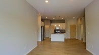This single-floor space features a thoughtfully designed open-concept layout that integrates the living area and kitchen into one cohesive environment. The light-toned hardwood flooring runs throughout, contributing to a clean, contemporary aesthetic and visually expanding the space.

The kitchen is a central highlight, anchored by a white island with a dark countertop that provides both prep space and a casual dining surface. White cabinetry lines the upper and lower walls, complemented by stainless steel appliances, including a refrigerator, range, and microwave. Pendant lighting hangs above the island, adding a decorative touch alongside recessed ceiling lighting that illuminates the entire area evenly.

The open living area adjacent to the kitchen offers ample room for furniture arrangement, with neutral gray walls providing a versatile backdrop. A built-in storage or pantry unit is visible near the refrigerator, adding practical functionality. A warm wood-toned entry door is positioned near the kitchen, offering a welcoming transition point into the space. Overall, the unit presents a bright, functional, and modern environment.