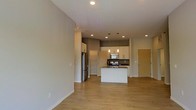 This single-floor space features a thoughtfully designed open-concept layout that integrates the living area and kitchen into one cohesive environment. The light-toned hardwood flooring runs throughout, contributing to a clean, contemporary aesthetic and visually expanding the space.

The kitchen is a central highlight, anchored by a white island with a dark countertop that provides both prep space and a casual dining surface. White cabinetry lines the upper and lower walls, complemented by stainless steel appliances, including a refrigerator, range, and microwave. Pendant lighting hangs above the island, adding a decorative touch alongside recessed ceiling lighting that illuminates the entire area evenly.

The open living area adjacent to the kitchen offers ample room for furniture arrangement, with neutral gray walls providing a versatile backdrop. A built-in storage or pantry unit is visible near the refrigerator, adding practical functionality. A warm wood-toned entry door is positioned near the kitchen, offering a welcoming transition point into the space. Overall, the unit presents a bright, functional, and modern environment.
