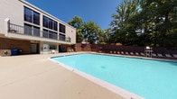 This single-floor residence combines functional design with distinctive character throughout its interior and outdoor spaces. Inside, the unit features well-appointed storage areas, including a built-in shelving unit with multiple tiers ideal for organizing everyday items, as well as a dedicated storage closet with carpeted flooring — spacious enough to accommodate sporting equipment such as a golf bag and additional storage boxes. A standout decorative element is the vintage-style bicycle wall art, a striking metalwork piece mounted on the interior walls that adds an artistic, industrial touch to the neutral-toned spaces. The interior walls are finished in soft, warm whites, contributing to a clean and airy atmosphere throughout. Beyond the interior, the property offers access to a generously sized outdoor swimming pool, which serves as a central outdoor amenity. The pool area is surrounded by a wide concrete deck furnished with lounge chairs and a shaded umbrella seating area, making it well-suited for relaxation. Mature trees frame the perimeter of the pool area, providing natural shade and greenery against the open blue sky. The adjacent multi-story building features large dark-tinted windows and balconies that overlook the pool, adding an architectural layer to the outdoor setting.