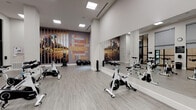 Located at 2629 Wagon Wheel Rd in Oxnard, CA, this dedicated spin studio is a well-appointed fitness space designed to deliver an energizing and focused cycling experience. The studio features a fleet of professional-grade Spinning bikes arranged across a durable, wood-plank-style floor that lends the space a clean, contemporary aesthetic.

The room is defined by its soaring ceilings and bright, recessed panel lighting that ensures consistent, glare-free illumination throughout. A full-length wall mirror runs along one side, visually expanding the space and allowing riders to monitor their form during workouts. On the opposing wall, a bold and vibrant motivational word-art mural — featuring terms like "Strength," "Discipline," "Balance," and "Energy" — serves as a dynamic focal point and sets an energizing tone for the room.

Natural light filters in through tall, black-framed windows fitted with roller shades, offering a connection to the lush outdoor surroundings while maintaining adjustable privacy. Additional amenities include a wall-mounted flat-screen display, stability balls, resistance bands, and a weight rack, supporting a variety of supplemental training activities. A wall clock and posted informational signage add functional touches to the space. The studio's modern design, thoughtful layout, and motivational atmosphere make it a well-equipped destination for cycling and group fitness.