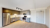 This single-floor unit presents a thoughtfully designed layout with modern finishes and a clean, contemporary aesthetic. The kitchen serves as a central highlight, featuring an L-shaped configuration with gray flat-front cabinetry extending from floor to ceiling, providing generous storage throughout. White countertops offer ample workspace, complemented by a full suite of stainless steel appliances including a range, over-the-range microwave, dishwasher, and refrigerator. A white subway tile backsplash adds a crisp, polished backdrop, while light hardwood-style flooring runs throughout the space, adding warmth. Black track lighting overhead provides directed, adjustable illumination across the kitchen and adjoining open area.

The bedroom is bright and airy, benefiting from a floor-to-ceiling window that frames city views and allows abundant natural light to fill the room. Neutral wall tones keep the space feeling calm and versatile.

The bathroom features a blue vanity with a white countertop and undermount sink, paired with a round backlit mirror for a modern touch. A full tub and shower combination with white subway tile surround and brushed nickel fixtures completes the space. Light hardwood-style flooring ties the bathroom's design cohesively to the rest of the unit.