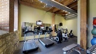 This fitness center combines a functional workout environment with distinctive industrial-inspired architecture. The space is defined by its striking exposed wooden beams and large cylindrical ductwork running along the open ceiling, lending a raw, warehouse-style character to the room. Recessed and track lighting ensures the space is well-lit throughout.

The facility is outfitted with a comprehensive selection of modern cardiovascular equipment, including multiple treadmills, elliptical trainers, and stationary bikes, all arranged to maximize floor space and ease of movement. A set of free weights and a weight bench are also available for strength training. Wall-to-wall mirrors along one side of the room visually expand the space and allow users to monitor their form during workouts.

Mounted televisions are positioned for easy viewing from the cardio machines, enhancing the exercise experience. The flooring consists of durable, rubber-textured material well-suited for a high-traffic fitness environment. The warm, sage-toned walls complement the natural wood elements overhead, creating an inviting atmosphere that balances the industrial aesthetic with a welcoming workout setting. Overall, the space is thoughtfully designed to support a complete fitness routine in a visually engaging environment.
