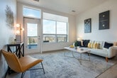 This contemporary single-floor residence showcases a thoughtfully designed open-concept layout that seamlessly connects the kitchen, dining, and living areas. The kitchen is outfitted with dark wood cabinetry, open shelving, stainless steel appliances including a French door refrigerator and electric range, a textured tile backsplash, and a generous island with a light quartz countertop and seating for bar-height chairs. Light wood-tone flooring runs throughout, adding warmth to the crisp white walls and ceilings.

The living area is anchored by oversized windows and a glass door that flood the space with natural light while offering elevated views of the surrounding landscape and rooftops. The bright, airy atmosphere is further enhanced by the open sightlines from the kitchen through to the living room.

The bedroom is a calm, well-proportioned retreat featuring a large picture window with outdoor views, a dedicated closet with hanging space, and direct access to the kitchen area. The bathroom is clean and functional, featuring light wood flooring, a glass-enclosed shower, and a minimalist white finish throughout.

Overall, the unit presents a modern, cohesive aesthetic defined by natural light, neutral tones, and practical design details.