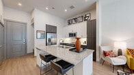 Located at 2650 S McDonald St in McKinney, TX, this thoughtfully designed one-bedroom, one-bathroom residence offers a contemporary living experience across a single floor. The open-concept layout seamlessly connects the kitchen, dining, and living areas, creating a bright and airy atmosphere enhanced by high ceilings and large windows dressed with patterned curtains and blinds that invite natural light throughout.

The kitchen is a standout feature, showcasing a granite-topped island with built-in shelving and bar seating, crisp white upper cabinetry contrasted by dark lower cabinets, a herringbone tile backsplash, and a full suite of stainless steel appliances including a French door refrigerator, range, microwave, and dishwasher. Light wood-tone flooring flows throughout the living areas, contributing to the home's modern, cohesive aesthetic.

The bedroom is a tranquil retreat with generous proportions and large windows, while the bathroom features granite countertops, a dark wood vanity, and a tub-shower combination with tile surround. Direct access from the bathroom leads to a well-organized walk-in closet with dual hanging rods and shelving. A dedicated laundry area and additional closet space round out this efficiently designed residence.