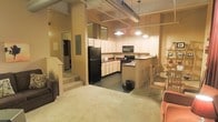 This single-floor space showcases an industrial loft aesthetic, highlighted by exposed ductwork and structural columns running along the ceiling, lending the interior a distinctly urban character. The open-concept layout allows the kitchen, dining, and living areas to flow seamlessly into one another, creating a spacious and connected environment.

The kitchen is well-equipped, featuring black appliances including a full-size refrigerator, a range with a stovetop, and an overhead microwave. White cabinetry lines the walls, providing ample storage, while a peninsula counter extends outward to define the kitchen space and offer bar-style seating with accompanying stools. Pendant lighting hangs above, adding warm illumination to the area.

Adjacent to the kitchen, a dining area accommodates a table and chairs, while the carpeted living area provides a comfortable retreat. A staircase visible in the background suggests access to a lower level or entry area. The combination of industrial ceiling details, practical kitchen features, and open floor planning gives this space a functional yet characterful feel suited to everyday living.