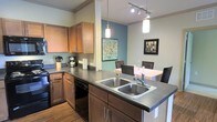This single-floor residence offers a thoughtfully designed interior that balances functionality with modern style. The kitchen serves as a central highlight, featuring rich warm-toned wood cabinetry, dark laminate countertops, and a double stainless steel sink set into a peninsula that also acts as a natural divider between the kitchen and dining area. Black appliances, including a dishwasher and range, complement the cabinetry and contribute to a cohesive, contemporary aesthetic. Overhead, a combination of pendant lighting and a track lighting system provides both ambient and task illumination, enhancing the kitchen's practicality and visual appeal. Crown molding along the ceiling adds a refined architectural touch throughout the space. The dining area is defined by a teal accent wall that introduces a pop of color, creating a warm and inviting atmosphere. Framed artwork adorns the walls, adding personality and character. The open-concept layout encourages easy interaction between cooking and dining areas, making the space well-suited for both everyday living and entertaining. Durable wood-look flooring grounds the interior with a warm, natural feel that ties the overall design together.