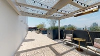 This expansive rooftop terrace offers a well-appointed outdoor retreat designed for both relaxation and entertaining. The space is anchored by a striking white pergola structure fitted with decorative geometric lattice panels that cast intricate patterned shadows across the light-toned pavers below. Strands of globe lights are draped throughout the pergola, adding ambiance for evening use, while ceiling-mounted heaters extend year-round comfort.

Beneath the pergola, a fully equipped outdoor kitchen features a stainless steel grill, countertop workspace, and a sink, complemented by a mounted flat-screen television and ample dining seating nearby. A separate lounge zone is centered around a rectangular fire pit with a glass wind guard, surrounded by generous blue sectional sofas and accent chairs, creating an ideal gathering area with unobstructed city skyline views.

Large dark metal planters filled with ornamental grasses, succulents, flowering shrubs, and small trees are strategically positioned throughout, adding greenery and natural texture to the urban setting. Additional seating clusters with gold-accented side tables and cushioned chairs provide intimate spaces under the pergola. Glass-and-aluminum entry doors connect the terrace to the building interior, completing this thoughtfully designed outdoor space.