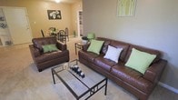 This single-floor residence offers a comfortable and cohesive living environment designed with everyday functionality in mind. The living area serves as the heart of the space, furnished with rich brown leather sofas accented by green and patterned throw pillows, creating a warm and inviting atmosphere. A glass-top coffee table with a metal frame anchors the seating arrangement over a light-toned carpet, adding a contemporary touch to the room.

The open layout allows for a seamless flow between the living area and the dining space, which is visible in the background and features a dark-framed dining table with matching chairs. This openness enhances the sense of space and makes the area feel airy and connected. Warm beige and taupe wall tones run throughout, contributing to a calm and neutral ambiance.

A built-in shelving unit is visible along the back wall, offering both storage and display opportunities. Decorative artwork, including a floral canvas and a nature-inspired print, adds personality to the walls. Accent lamps with green shades complement the décor and provide additional lighting. The overall design reflects a balanced blend of comfort and style, making this a welcoming and practical living environment.