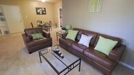 This single-floor residence offers a comfortable and cohesive living environment designed with everyday functionality in mind. The living area serves as the heart of the space, furnished with rich brown leather sofas accented by green and patterned throw pillows, creating a warm and inviting atmosphere. A glass-top coffee table with a metal frame anchors the seating arrangement over a light-toned carpet, adding a contemporary touch to the room.

The open layout allows for a seamless flow between the living area and the dining space, which is visible in the background and features a dark-framed dining table with matching chairs. This openness enhances the sense of space and makes the area feel airy and connected. Warm beige and taupe wall tones run throughout, contributing to a calm and neutral ambiance.

A built-in shelving unit is visible along the back wall, offering both storage and display opportunities. Decorative artwork, including a floral canvas and a nature-inspired print, adds personality to the walls. Accent lamps with green shades complement the décor and provide additional lighting. The overall design reflects a balanced blend of comfort and style, making this a welcoming and practical living environment.