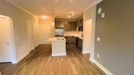 This single-floor unit showcases an open-concept layout that seamlessly connects the kitchen and living areas, creating a spacious and functional environment. The kitchen is a standout feature, equipped with stainless steel appliances, including a refrigerator, microwave, and range. Dark taupe cabinetry with clean lines is complemented by light-toned countertops and a classic subway tile backsplash, striking a balance between contemporary and timeless design.

A central kitchen island with a white finish and an integrated sink serves as both a practical workspace and a natural gathering point. Overhead, a decorative pendant light adds a warm, stylish accent above the island, while track lighting illuminates the cooking area. Consistent hardwood-style flooring flows throughout the space, adding warmth and visual continuity.

Crown molding along the ceiling lends a refined architectural detail to the interior. The open layout allows natural sightlines across the space, enhancing the sense of openness. Neutral gray and white wall tones provide a versatile backdrop, while multiple doorways suggest easy access to additional rooms and storage areas within the unit.