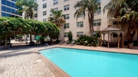 This amenity space offers residents two distinct areas designed for relaxation and fitness. The outdoor pool area is set within a courtyard framed by a multi-story building, lush tropical palms, and abundant greenery. The sparkling turquoise pool is surrounded by a brick-paved deck, lounge chairs for sunbathing, and a covered gazebo, creating a serene retreat ideal for unwinding outdoors.

Adjacent to the pool, the indoor fitness center is a generously sized, well-lit space featuring large windows that invite ample natural light and offer views of the surrounding property. The room is finished with stylish herringbone-patterned carpet flooring and is equipped with an impressive array of exercise equipment. Strength training options include a power rack, cable machine, dumbbell rack with a full range of free weights, kettlebells, medicine balls, battle ropes, and resistance bands. Cardio equipment includes treadmills and a rowing machine, while a Precor stationary bike rounds out the offerings. Full-length mirrors along one wall expand the sense of space, and ceiling fans provide comfortable air circulation. A wall-mounted television near the cardio area adds entertainment during workouts, making this fitness center a thorough and functional facility.