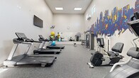 Located at 9001 Renner Blvd in Lenexa, Kansas, this fitness center offers a thoughtfully designed single-room workout space with a generous open floor plan. The room features durable rubber flooring, crisp white walls, and elevated ceilings that lend an airy feel to the environment. Natural light filters in through high clerestory windows, complemented by recessed panel lighting overhead for consistent, even illumination throughout.

A striking abstract cityscape mural spans one wall, adding bold color and visual energy to the space. A large wall-mounted mirror runs along another wall, enhancing the sense of depth and allowing users to monitor their form during exercise. A wall-mounted flat-screen television and an analog clock provide practical amenities for workout sessions.

The fitness center is well-stocked with a variety of equipment. Cardio options include multiple Spirit-brand treadmills and elliptical machines, as well as a stationary bike. Strength training is supported by a multi-function cable machine, a dumbbell rack with a full range of weights, a workout bench, and a medicine ball rack with an assortment of exercise balls in various sizes. A wall-mounted fan provides additional air circulation, alongside a dedicated HVAC unit. The layout provides ample floor space between equipment, allowing for comfortable, unobstructed movement throughout the room.