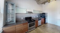 Located on Beverly Boulevard in the heart of Los Angeles, this single-floor space showcases a compelling industrial aesthetic that blends raw architectural character with functional modern updates. Exposed wood beam ceilings and polished concrete floors run throughout the open main area, creating a loft-like atmosphere with generous ceiling height and a sense of expansive volume. Structural concrete columns add to the industrial character of the main living area, which benefits from multiple windows that invite natural light and offer views of the surrounding neighborhood.

The kitchen is thoughtfully appointed with warm-toned wood cabinetry, sleek grey countertops, frosted glass upper cabinets, a stainless steel range, dishwasher, and refrigerator — all set against a stainless steel backsplash and complemented by a ventilation hood. Additional rooms feature the same polished concrete flooring with bright white walls and windows providing natural light and outdoor views. The bathroom stands out with bold black marble-style tile surrounding the soaking tub and shower, classic white hex mosaic floor tile, and a window framing views of swaying palm trees — a quintessentially Los Angeles detail that ties the entire space together.