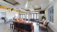 This single-floor community space combines comfort and style in a well-appointed layout designed for gathering and relaxation. The main lounge area is anchored by a set of large, multi-pane French doors that flood the room with natural light and provide an open, airy atmosphere. Coffered ceilings with recessed lighting add architectural depth and elegance overhead, while a wall-mounted flat-screen television offers entertainment for residents and guests. Comfortable seating, including plush orange sofas and accent chairs, is arranged around a central coffee table, creating an inviting conversation area. Herringbone-patterned tile flooring runs throughout, lending a refined, contemporary touch to the space. Adjacent to the lounge, a kitchenette is outfitted with dark wood cabinetry, granite-style countertops, a sink, and a microwave, making it ideal for hosting events or casual gatherings. Warm wood-paneled ceiling accents and pendant chandelier lighting in the kitchenette zone add a rustic charm that complements the overall design. A glimpse into an adjoining fitness area is visible through a mirrored wall, rounding out the suite of amenities. The space balances functionality with an inviting aesthetic throughout.