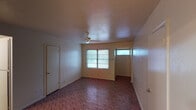 This single-floor unit offers a practical and straightforward layout across several rooms, all unified by distinctive parquet-patterned flooring. The main living area is the most spacious room in the unit, featuring a ceiling fan, a textured ceiling, and windows that allow natural light to enter while providing views to the outdoors. A front entry door with a transom window above adds additional light to the entryway.

The unit includes multiple additional rooms that can serve as bedrooms or flexible-use spaces. Each room features the same parquet flooring, textured ceilings, and windows fitted with blinds. One room benefits from dual windows on adjacent walls, allowing for cross-ventilation and generous natural light.

The kitchen is equipped with white cabinetry extending to the ceiling, a gas range with overhead ventilation, a dishwasher, a full-size refrigerator, and a sink with a window above. The kitchen flooring continues the parquet theme seen throughout the unit.

The bathroom includes a full tub and shower combination with white tile surround, a toilet, and a small window for ventilation. Overall, the unit presents a functional, consistent interior with neutral tones throughout.