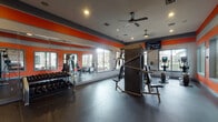 This single-floor fitness center is designed to deliver a complete workout experience in a vibrant, energizing environment. The interior is defined by a striking color scheme of bold orange accent walls contrasted against gray trim and a white ceiling, creating a dynamic, motivating atmosphere. Durable rubber flooring spans the entire space, providing both safety and comfort during exercise.

The main workout area is generously proportioned, offering ample room to move freely between equipment stations. A full-length wall mirror along one side visually expands the space and allows users to monitor their form during training. Multiple ceiling fans ensure continuous air circulation, enhancing comfort throughout workouts.

Large windows line the walls on multiple sides, flooding the interior with natural light and providing views of the surrounding outdoor area, including what appears to be a covered patio or courtyard. This connection to the outdoors contributes to an open, airy feel.

Equipment includes a multi-station cable machine, a rack of free weights ranging across various sizes, and several cardio machines including an upright bike and elliptical trainer. A wall-mounted flat-screen television is positioned for visibility throughout the space. The fitness center is accessed through a glass-paneled wooden door, with posted fitness center rules clearly displayed at the entrance.