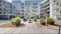 Located at 1350 Speer Blvd in Denver, Colorado, this expansive outdoor courtyard serves as a central communal gathering space surrounded by a modern multi-story building with balconied residences on all sides. The courtyard is thoughtfully designed to encourage social interaction and outdoor living, with several distinct activity zones connected by wide concrete paver pathways.

At the heart of the space, a comfortable lounge area features stylish outdoor seating arranged around a central fire pit table, framed by large decorative planters filled with ornamental grasses and colorful seasonal flowers. Gravel landscaping beds add texture and low-maintenance greenery throughout.

A contemporary pergola structure with slatted steel louvers and strung bistro lighting anchors the dining zone, sheltering a long communal dining table with ample seating — ideal for group gatherings. Adjacent to this area is a built-in outdoor kitchen equipped with a stainless steel grill and dark granite countertops.

A full-length bocce ball court provides an additional recreational amenity, adding a lively, social dimension to the courtyard. Smaller seating alcoves with wicker lounge chairs are tucked along the building's edges, offering quieter retreats. The open-sky design floods the entire courtyard with natural light throughout the day.