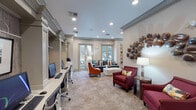 This single-floor amenity space is designed to serve both productivity and comfort, blending professional functionality with refined interior design. The main lounge area features plush seating, including a teal velvet sofa, vibrant orange accent chairs, and a sleek white coffee table, all set atop patterned carpeting. Large windows along the perimeter flood the space with natural light and offer views of lush outdoor greenery, enhancing the welcoming ambiance. Crown molding and recessed lighting throughout lend an upscale, polished character to the interiors. A built-in workstation area lines one wall, featuring multiple computer terminals framed by decorative columns and textured wallcovering, with framed artwork displayed above each station. Adjacent to the main lounge, a dedicated conference room provides a more intimate setting for meetings, centered around a substantial granite-topped table surrounded by ergonomic chairs. Geometric pendant lighting and large-scale photographic artwork of river stones add visual interest to this room. Decorative wall art, including a sculptural metallic piece and a mosaic-style wood panel mirror, add distinctive character throughout the space. The overall atmosphere is sophisticated yet inviting, making it well-suited for both focused work and collaborative gatherings.