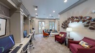 This single-floor amenity space is designed to serve both productivity and comfort, blending professional functionality with refined interior design. The main lounge area features plush seating, including a teal velvet sofa, vibrant orange accent chairs, and a sleek white coffee table, all set atop patterned carpeting. Large windows along the perimeter flood the space with natural light and offer views of lush outdoor greenery, enhancing the welcoming ambiance. Crown molding and recessed lighting throughout lend an upscale, polished character to the interiors. A built-in workstation area lines one wall, featuring multiple computer terminals framed by decorative columns and textured wallcovering, with framed artwork displayed above each station. Adjacent to the main lounge, a dedicated conference room provides a more intimate setting for meetings, centered around a substantial granite-topped table surrounded by ergonomic chairs. Geometric pendant lighting and large-scale photographic artwork of river stones add visual interest to this room. Decorative wall art, including a sculptural metallic piece and a mosaic-style wood panel mirror, add distinctive character throughout the space. The overall atmosphere is sophisticated yet inviting, making it well-suited for both focused work and collaborative gatherings.