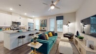 This thoughtfully designed single-floor residence blends modern aesthetics with functional living. The open-concept layout seamlessly connects the kitchen and living area, creating an inviting, airy atmosphere. The kitchen is outfitted with white shaker cabinetry, granite countertops, a large peninsula with seating, stainless steel appliances including a French-door refrigerator, microwave, and range, and elegant pendant lighting overhead. Wood-look flooring runs throughout, adding warmth and continuity across the space. The living room is generously proportioned, featuring a ceiling fan, large windows with blinds that invite natural light, and ample room for comfortable seating arrangements. A hallway lined with built-in open shelving provides convenient storage and leads to the private quarters. The bedroom is a serene retreat, highlighted by a large window with teal drapery, offering views of greenery outside. It connects directly to a full bathroom, which includes a vanity with white cabinetry, a bathtub with a teal shower curtain, and wood-look flooring. Neutral wall tones and clean architectural lines give the entire space a cohesive, contemporary character, while teal and gold accent tones add a vibrant, curated touch throughout.