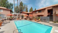 This inviting outdoor recreational area is centered around a generously sized, sparkling swimming pool with crystal-clear blue water, ideal for leisure and relaxation. The pool deck is lined with lounge chairs perfect for sunbathing, while a shaded patio area nearby features tables and chairs beneath vibrant orange umbrellas, offering a comfortable spot to unwind outdoors. A separate hot tub spa is conveniently located adjacent to the main pool area, providing an additional relaxation option. An outdoor rinse shower is positioned near the pool entrance, allowing guests to freshen up with ease. The surrounding grounds are landscaped with potted trees and desert-adapted plants, complementing the warm, earthy tones of the Spanish-influenced architecture. The buildings feature a stucco exterior with terracotta-tiled rooflines and multi-level balconies, framing the courtyard with a classic Southwestern aesthetic. Tall palm and pine trees rise above the rooftops, providing a lush natural backdrop and partial shade. Safety signage is prominently displayed throughout the area. The entire pool enclosure is secured with metal fencing, ensuring a safe and well-maintained environment for all users.
