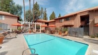 This inviting outdoor recreational area is centered around a generously sized, sparkling swimming pool with crystal-clear blue water, ideal for leisure and relaxation. The pool deck is lined with lounge chairs perfect for sunbathing, while a shaded patio area nearby features tables and chairs beneath vibrant orange umbrellas, offering a comfortable spot to unwind outdoors. A separate hot tub spa is conveniently located adjacent to the main pool area, providing an additional relaxation option. An outdoor rinse shower is positioned near the pool entrance, allowing guests to freshen up with ease. The surrounding grounds are landscaped with potted trees and desert-adapted plants, complementing the warm, earthy tones of the Spanish-influenced architecture. The buildings feature a stucco exterior with terracotta-tiled rooflines and multi-level balconies, framing the courtyard with a classic Southwestern aesthetic. Tall palm and pine trees rise above the rooftops, providing a lush natural backdrop and partial shade. Safety signage is prominently displayed throughout the area. The entire pool enclosure is secured with metal fencing, ensuring a safe and well-maintained environment for all users.