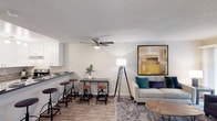 This single-floor residential unit offers a thoughtfully designed layout that balances comfort and functionality. The exterior presents a multi-story building facade with mature trees and a private vinyl privacy fence enclosing an outdoor area with decorative ground cover, offering a sense of seclusion.

Inside, the open-concept living and dining area features wood-look flooring, neutral walls, and a textured ceiling. A ceiling fan provides overhead comfort, while a sliding glass door floods the space with natural light and provides direct access to the fenced patio. The kitchen is outfitted with white shaker-style cabinetry, dark countertops, a mosaic tile backsplash, stainless steel appliances including a refrigerator and range, and a breakfast bar with seating.

The bedrooms are carpeted and finished with soft neutral tones, featuring window blinds that allow natural light while maintaining privacy. Ample closet space with sliding doors is present in the primary bedroom. The bathrooms are clean and functional, featuring dark countertops, white cabinetry, large mirrors, and well-placed lighting. A hallway connects the living spaces to the private quarters, completing an efficient and comfortable floor plan.
