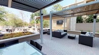 This expansive outdoor amenity space in Dallas is thoughtfully designed to offer a refined al fresco experience across a single level. The centerpiece of the space is a striking circular water feature surrounded by river rock and flagstone paving, encircled by wicker lounge chairs that invite relaxation. Multiple distinct zones provide varied settings for gathering and leisure. A sleek modern pergola with slatted overhead panels creates a shaded outdoor living area, complete with contemporary wicker sectional sofas and a wall-mounted television — ideal for entertaining. Beneath a separate pergola structure, a large glass-topped dining table with wicker chairs offers an elegant setting for outdoor meals. A manicured lawn area provides open green space alongside a wood-deck terrace furnished with round bistro tables and chairs. Raised planters and lush landscaping with mature trees add natural texture throughout, while cable-rail fencing preserves open sightlines to the surrounding Dallas skyline and tree canopy. Large floor-to-ceiling glass sliding doors connect the outdoor terrace to an interior space, seamlessly blending indoor and outdoor living. The brick-paved courtyard entry is framed by well-maintained hedgerows and multiple glass-fronted building entries, reinforcing the polished, community-oriented character of this urban outdoor retreat.