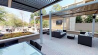 This expansive outdoor amenity space in Dallas is thoughtfully designed to offer a refined al fresco experience across a single level. The centerpiece of the space is a striking circular water feature surrounded by river rock and flagstone paving, encircled by wicker lounge chairs that invite relaxation. Multiple distinct zones provide varied settings for gathering and leisure. A sleek modern pergola with slatted overhead panels creates a shaded outdoor living area, complete with contemporary wicker sectional sofas and a wall-mounted television — ideal for entertaining. Beneath a separate pergola structure, a large glass-topped dining table with wicker chairs offers an elegant setting for outdoor meals. A manicured lawn area provides open green space alongside a wood-deck terrace furnished with round bistro tables and chairs. Raised planters and lush landscaping with mature trees add natural texture throughout, while cable-rail fencing preserves open sightlines to the surrounding Dallas skyline and tree canopy. Large floor-to-ceiling glass sliding doors connect the outdoor terrace to an interior space, seamlessly blending indoor and outdoor living. The brick-paved courtyard entry is framed by well-maintained hedgerows and multiple glass-fronted building entries, reinforcing the polished, community-oriented character of this urban outdoor retreat.
