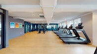 This modern fitness center is designed to support a comprehensive workout experience across a single, open floor. The space is finished with light hardwood flooring throughout, creating a clean and energetic atmosphere complemented by bold teal and slate-blue accent walls. Recessed lighting and exposed ductwork along the ceiling contribute to the facility's contemporary aesthetic.

The cardio zone is generously outfitted with multiple treadmills, elliptical trainers, and stationary bikes, all positioned along large windows that allow abundant natural light and offer views of the surrounding cityscape. Full-length mirrors line portions of the walls, enhancing the sense of openness and allowing users to monitor their form.

A dedicated strength training area features a multi-station cable machine and a variety of resistance equipment, providing options for a wide range of exercises. An adjacent open floor area, framed by glass partition walls, serves as a flexible studio space suitable for stretching, cycling, or group activities.

Motivational fitness posters are displayed on the walls, adding an encouraging touch to the environment. The facility's layout is thoughtfully organized to keep cardio and strength zones distinct while maintaining an open, uncluttered flow throughout the space.