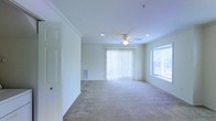 This two-floor residential space offers a clean, well-maintained interior designed for comfortable everyday living. The main living area is generously sized, featuring soft wall-to-wall carpeting that adds warmth underfoot and a neutral color palette throughout that lends itself to a variety of décor styles. Crown molding accents the ceilings, adding a refined architectural detail that elevates the overall finish of the space.

Natural light plays a significant role in the ambiance of the living area, with large windows fitted with blinds and a set of sliding glass doors dressed in vertical blinds that open to an outdoor area. This abundance of glazing ensures the room feels bright and airy throughout the day. Recessed lighting and a centrally mounted ceiling fan with a light fixture provide functional illumination and climate comfort year-round.

A wall-mounted HVAC vent is also visible, indicating a built-in climate control system. The layout flows openly, offering flexible arrangement possibilities for furnishings. Spanning two floors, the overall footprint of this space provides ample room for both living and private areas, making it a practical and inviting place to call home.