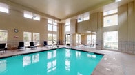 The pool area offers two distinct aquatic environments designed for year-round enjoyment. The indoor pool facility is an architecturally impressive space featuring soaring ceilings and an abundance of natural light streaming through rows of clerestory windows and large wall-mounted windows on multiple sides. The interior is finished with neutral toned walls and tile accents, creating a bright and welcoming atmosphere. The generously sized rectangular pool with vivid turquoise water is complemented by an adjacent hot tub, ideal for relaxation. Pool equipment and recreational accessories are conveniently available, and lounge chairs are positioned along the pool deck for comfort. A water fountain and direct exterior access are also provided within the space. The outdoor pool area presents a large, well-maintained swimming pool alongside a separate spa, both set within a spacious concrete deck lined with numerous lounge chairs and shaded umbrella tables. The outdoor setting is framed by mature trees and lush landscaping, offering a pleasant natural backdrop. Access to the outdoor pool is provided through a covered entry with a posted pool rules sign, ensuring an organized and enjoyable experience for all users.