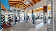 Located at 14435 South 48th Street in Phoenix, AZ, this expansive single-floor fitness center offers an impressive and thoughtfully designed exercise environment. The space is defined by its dramatic vaulted ceilings with exposed wooden beams and warm tongue-and-groove wood paneling, creating an inviting yet energizing atmosphere. Ceiling fans are strategically placed throughout to complement the climate control system.

Large floor-to-ceiling windows and glass wall panels flood the interior with abundant natural light while offering views of the outdoor surroundings, including palm trees and a pool area. The flooring transitions between wood-look planks and durable carpet tile, delineating functional workout zones throughout the space.

The exercise room is comprehensively outfitted with a wide selection of Matrix brand cardio equipment, including treadmills, ellipticals, and stair climbers arranged along the windowed perimeter. A dedicated strength training zone features multiple cable machines, weight racks, benches, medicine balls, and free weights. Full-length mirrors maximize the sense of space and support proper form during workouts.

Bold accent walls in vivid blue and orange add a dynamic visual energy to the space. A separate spin studio with stationary bikes is enclosed behind glass near the entrance. Stone veneer columns and architectural archways contribute refined structural character throughout this well-appointed fitness facility.