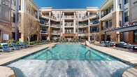 Located in the vibrant Dilworth neighborhood of Charlotte, NC, this outdoor amenity space is centered around a generously sized, resort-style swimming pool that serves as the focal point of a well-designed courtyard. The pool features a built-in wading entry and is flanked by an expansive concrete sundeck lined with lounge chairs and umbrella-shaded seating, ideal for relaxing under the Carolina sun.

The courtyard is enclosed on multiple sides by a striking multi-story building that blends classic brick masonry with contemporary gray siding and large-paned windows, lending the space an upscale yet welcoming ambiance. Private balconies on each floor overlook the pool area, creating a lively, connected atmosphere.

On the perimeter of the deck, a pergola-covered outdoor lounge area provides a shaded retreat, complemented by additional seating nooks bordered by decorative brick walls with built-in planters. The patio furniture throughout includes modern tables and chairs suited for both casual dining and socializing. Mature trees and manicured greenery soften the hardscape, adding a natural element to the urban setting.

The overall design of this outdoor space balances functionality with aesthetic appeal, offering a polished and inviting environment that reflects the quality and character of its South End Charlotte location.