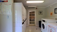 This shared laundry facility is designed with practicality and convenience in mind. The room features a row of stacked washer and dryer units lining the left wall, offering ample machine availability for multiple users. On the right side, a front-load washing machine provides additional capacity, complemented by a large waste bin nearby for easy disposal. The space is finished with neutral gray walls and light-toned flooring, creating a clean and functional atmosphere. Overhead, a flush fluorescent light fixture and an additional recessed vent panel ensure the room is well-lit and properly ventilated. A ceiling-mounted HVAC unit further contributes to a comfortable environment. A framed piece of artwork on the wall adds a subtle decorative touch, softening the utilitarian character of the room. A prominent interior door with a multi-pane glass grid allows natural light to filter in from the adjacent common area, which is visible through the window and appears to feature warm-toned walls and furnishings. The glazed door also provides a visual connection between spaces, enhancing the open and accessible feel of the facility. Overall, this laundry room is a well-maintained, thoughtfully arranged space that balances functionality with a tidy, welcoming presentation.