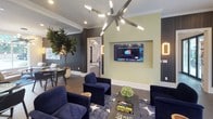 This thoughtfully designed single-floor amenity space combines a café, lounge, and outdoor connectivity into a cohesive, upscale environment. The Radius Café anchors one section of the space, featuring a stainless steel service counter with bar-height seating, a bold geometric patterned accent wall, and a retro-style lime green refrigerator that adds a playful pop of color. Glass pendant lights illuminate the counter area, while a built-in banquette with high upholstered backing and a round bistro table create an intimate dining nook nearby. Dark hardwood-style flooring flows throughout, grounding the warm, neutral palette. Two striking large-format abstract paintings with gold and charcoal tones serve as a visual focal point connecting the café to the lounge. The lounge area features navy velvet armchairs arranged around a geometric coffee table beneath a dramatic sputnik-style chandelier. A wall-mounted flat-screen TV is set into a recessed niche, flanked by dark wood panel accents and warm sconce lighting. A mature fiddle-leaf fig tree adds organic texture. French doors at the rear of the lounge open directly to an outdoor pool area, flooding the space with natural light and providing seamless indoor-outdoor access.