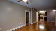 This single-floor residence offers a thoughtfully designed layout that balances comfort and functionality. The main living area is highlighted by rich, warm-toned hardwood flooring that extends throughout, adding a sense of elegance and continuity to the space. A ceiling fan is centrally mounted overhead, providing both practical airflow and a classic aesthetic touch. Neutral taupe-toned walls create a calm, versatile backdrop that complements the warm flooring and white trim work.

A doorway from the living area leads to an adjacent room, suggesting a flexible layout suited for varied living arrangements. The kitchen, visible from the main living space, features warm wood cabinetry with ample storage, light-toned countertops, and tile flooring that transitions naturally from the hardwood in the living area. The kitchen is well-lit with overhead lighting, giving it a bright and functional character.

White baseboards and clean door trim throughout the space add a crisp, finished look. The open sightlines between the living area and kitchen contribute to an airy, connected feel, making the space well-suited for everyday living. Overall, this residence combines practical design with warm, inviting finishes for a comfortable living environment.