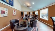This single-floor lounge space exudes a welcoming ambiance through its rich, earth-toned walls, warm hardwood flooring, and vaulted ceilings that lend a sense of openness and volume to the interior. The space is thoughtfully arranged into multiple seating zones, beginning with a central casual lounge area furnished with dark leather barrel chairs grouped around a round wooden coffee table, accented by a colorful floral centerpiece and anchored by a decorative area rug. A wall-mounted flat-screen television and music-themed artwork panels add personality and visual interest to the warm-toned walls. Toward the far end of the room, a raised bar-height table with ornately carved dark wood chairs provides a more intimate seating option, accompanied by potted decorative trees that introduce a touch of greenery. Professional speakers are positioned in this area, suggesting the space is equipped for live or amplified audio. Large windows along the side walls allow natural light to filter in, brightening the interior while offering views to the outside. Recessed lighting and a central ceiling fixture provide additional illumination throughout. A small service counter or refreshment station is also visible, adding functional convenience to the space. Overall, the room is designed to foster a relaxed, social atmosphere with a distinct musical character.
