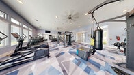 Located at 1520 Alexis Ave in Daytona Beach, FL, this fitness center offers a comprehensive workout environment spread across a single, generously sized floor. The main exercise room is a standout space, featuring striking geometric-patterned carpet in shades of blue, gray, and white that lends a dynamic, modern aesthetic. Ceiling fans and recessed lighting keep the area bright and comfortable, while a row of large windows along the walls floods the space with natural light and views of the surrounding landscape. The main floor is outfitted with an extensive array of equipment, including treadmills, cable machines, a multi-functional power rack, a punching bag, TRX suspension trainers, a plyo box, weight benches, and a full dumbbell rack — catering to cardio, strength, and functional training alike. A mounted flat-screen TV adds convenience for entertainment during workouts. Adjacent to the main floor, a separate studio room provides a quieter setting with light hardwood-style flooring, full-length mirrors, a wall-mounted TV, and equipment such as stationary bikes, a rowing machine, and stability balls — ideal for low-impact or focused training sessions. The hallway connects the two spaces seamlessly, completing this well-appointed, full-service fitness facility.