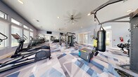 Located at 1520 Alexis Ave in Daytona Beach, FL, this fitness center offers a comprehensive workout environment spread across a single, generously sized floor. The main exercise room is a standout space, featuring striking geometric-patterned carpet in shades of blue, gray, and white that lends a dynamic, modern aesthetic. Ceiling fans and recessed lighting keep the area bright and comfortable, while a row of large windows along the walls floods the space with natural light and views of the surrounding landscape. The main floor is outfitted with an extensive array of equipment, including treadmills, cable machines, a multi-functional power rack, a punching bag, TRX suspension trainers, a plyo box, weight benches, and a full dumbbell rack — catering to cardio, strength, and functional training alike. A mounted flat-screen TV adds convenience for entertainment during workouts. Adjacent to the main floor, a separate studio room provides a quieter setting with light hardwood-style flooring, full-length mirrors, a wall-mounted TV, and equipment such as stationary bikes, a rowing machine, and stability balls — ideal for low-impact or focused training sessions. The hallway connects the two spaces seamlessly, completing this well-appointed, full-service fitness facility.