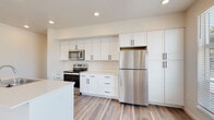 This single-floor unit presents a clean, contemporary design highlighted by consistent light-toned wood-look flooring and a neutral color palette that flows seamlessly throughout the space. The kitchen serves as a central feature, showcasing white shaker-style cabinetry with matte black hardware extending floor to ceiling for generous storage capacity. Stainless steel appliances, including a refrigerator, range, microwave, and dishwasher, complement the bright white countertops. A kitchen island provides additional workspace and sink access, while large windows allow abundant natural light to filter into the adjoining living area.

The bedrooms are well-proportioned, each featuring windows that bring in natural light and views of surrounding greenery. One bedroom includes a spacious walk-in closet fitted with wire shelving and hanging rods along multiple walls, offering practical organization. The full bathroom is finished with a white vanity, undermount sink, large wall mirror, a tub-shower combination, and matte black accent hardware that ties into the home's consistent design language. A dedicated entry area and in-unit laundry space complete the functional layout of this thoughtfully designed residence.