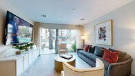 This single-floor space showcases a thoughtfully designed open-concept layout that seamlessly connects its key living areas. Throughout the space, light-toned herringbone-patterned flooring adds a refined architectural detail that ties each room together cohesively.

The living area is bright and welcoming, highlighted by large floor-to-ceiling sliding glass doors that flood the room with natural light and offer direct views of an outdoor pool area with palm trees. The space is anchored by a charcoal sectional sofa with bold red accent pillows, complemented by curved cream accent chairs, a gold drum coffee table, and a wall-mounted flat-screen TV resting above a sleek marble-topped console cabinet. Curated artwork adorns the walls, adding a gallery-like quality.

The kitchen is well-appointed with light beige cabinetry extending to the ceiling, marble-style backsplash, stainless steel appliances, and a generous island topped with a white marble surface. Vibrant blue velvet barstools provide stylish seating at the island, making it ideal for casual dining or entertaining.

The dining area features a round white dining table surrounded by modern white chairs, centered beneath an elegant tiered chandelier. Framed abstract artwork lines the walls, enhancing the sophisticated atmosphere. A glass door connects the space to an adjacent common area, further emphasizing its open and airy character.