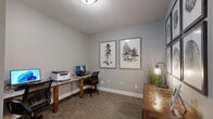Located at 100 Riverchase Boulevard in Nashville, Tennessee, this single-room office space offers a functional and thoughtfully designed work environment. The room is finished with neutral gray walls and soft carpeted flooring, creating a calm and professional atmosphere conducive to focused work.

A prominent feature of the space is the L-shaped granite-topped workstation, which provides ample surface area to accommodate multiple workstations, computer monitors, and office equipment such as a printer. Ergonomic task chairs complement the desk setup, making the space well-suited for productivity.

The room is accessed through a distinctive French door with a grid-pane window, adding an architectural detail that lends both style and a sense of openness. A decorative console table positioned against one wall is accented by a set of six framed botanical art prints arranged in a gallery-style grid, featuring tree ring motifs that lend a nature-inspired aesthetic to the space.

Additional wall art, including two framed watercolor tree prints, further reinforces the organic, natural theme throughout. Potted greenery and a sleek floor lamp add warmth and texture. Flush ceiling lighting ensures the room is well-illuminated. Overall, this office space balances practicality with an inviting, nature-inspired design sensibility.