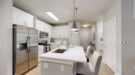 This single-floor residence showcases a clean, contemporary design throughout. The kitchen serves as a central highlight, featuring white cabinetry, a classic subway tile backsplash, quartz countertops, and a full suite of stainless steel appliances including a refrigerator, electric range, and built-in microwave. A generously sized island with an undermount sink provides additional prep space and seating for casual dining, accented by brushed nickel pendant lighting overhead. Light wood-tone flooring flows through the main living areas, enhancing the bright, airy atmosphere.

A dedicated dining nook accommodates a round table with seating for four, decorated with geometric gold-framed mirrors and wall art. The living area offers a comfortable, well-proportioned layout. The bedrooms are carpeted and receive natural light through large windows, creating a warm and restful environment.

Two bathrooms serve the residence: one featuring a glass-enclosed walk-in shower with white subway tile, and a second with a full soaking tub and tiled surround. Both bathrooms are finished with brushed nickel fixtures, maintaining a cohesive, modern aesthetic consistent with the rest of the home.