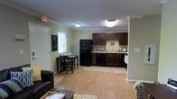 Located at 1110 E 10th St in Greenville, NC, this single-floor space offers a practical and welcoming open-concept layout that seamlessly connects the living, dining, and kitchen areas. The interior is finished with warm hardwood-style flooring that extends throughout the main living space, contributing to a cohesive and tidy aesthetic.

The kitchen is well-appointed with rich dark-stained cabinetry providing generous storage, light-toned countertops, and black appliances including a full-size refrigerator and range. The kitchen flows naturally into a dedicated dining area, which comfortably accommodates a small table and chairs — an ideal setup for everyday meals.

The living area anchors the open floor plan, offering space for comfortable seating and everyday relaxation. Crown molding along the ceiling adds a refined architectural detail that elevates the overall finish of the space. Natural light filters in through a window near the entryway, brightening the interior and contributing to an open, airy feel. The entry door features a welcoming touch with a small chalkboard sign, adding a personalized and homey character to the space. Ceiling-mounted light fixtures ensure consistent illumination throughout. Overall, the layout is efficient, functional, and move-in ready.